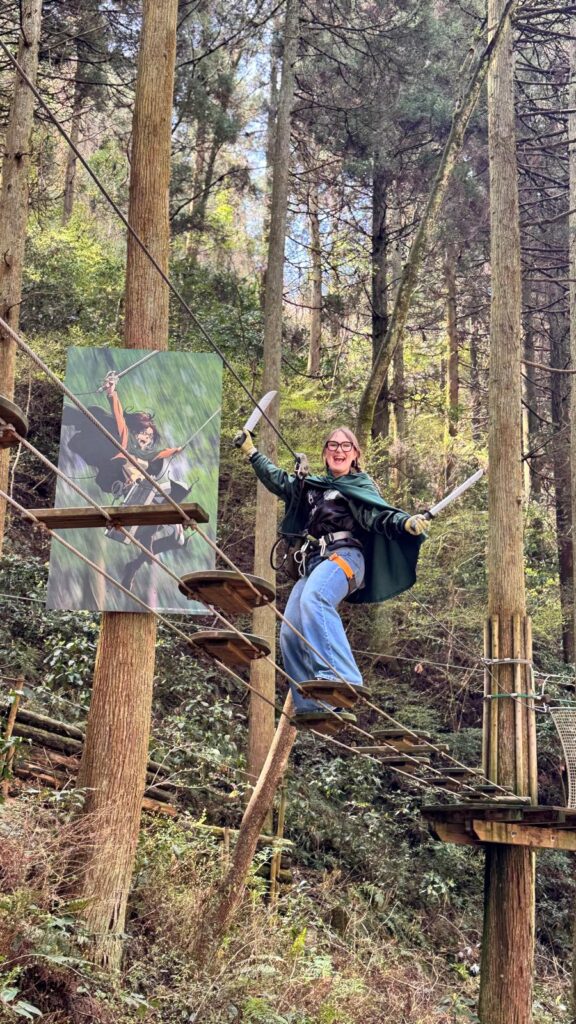 Persona sonriente cruza un puente colgante de cuerdas en medio de un bosque alto y frondoso. Lleva arnés de seguridad, guantes y una capa verde, y sostiene dos espadas de utilería en pose entusiasta. A su lado hay un cartel con un personaje de anime en una postura similar. El entorno natural está lleno de árboles altos y vegetación densa.