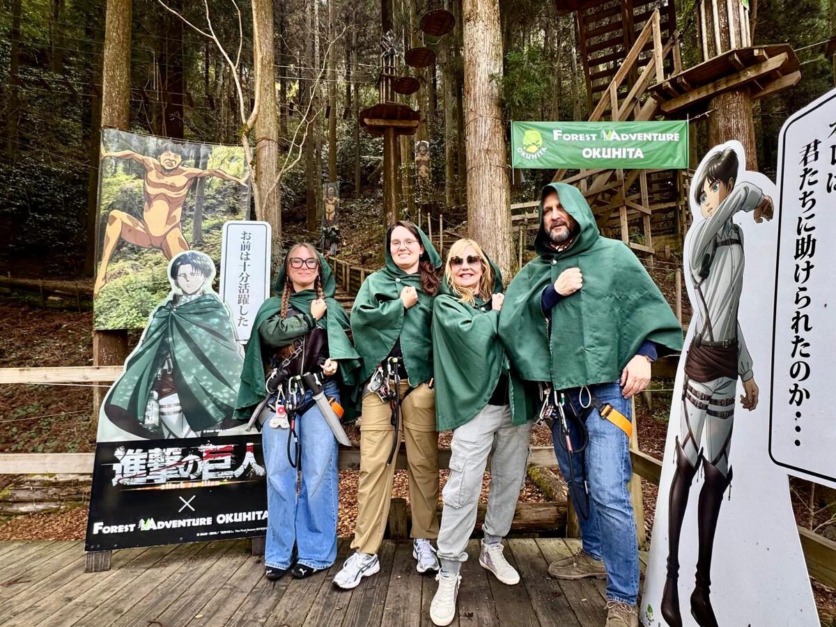 Cuatro personas posan juntas en una plataforma de madera en medio de un bosque, vestidas con capas verdes y arneses de seguridad, imitando a personajes de anime. A su alrededor hay carteles ilustrados con personajes y un letrero de “Forest Adventure Okuhita”.