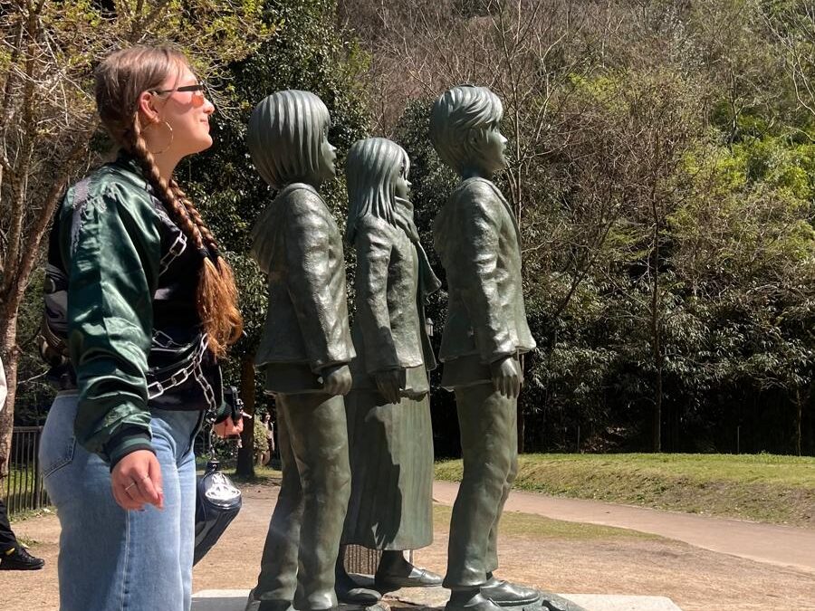 Persona de perfil observa una escultura de tres figuras humanas alineadas sobre un pedestal de piedra en un entorno natural. Lleva chaqueta verde, vaqueros y gafas de sol. Al fondo se ven árboles y una montaña bajo un cielo despejado.