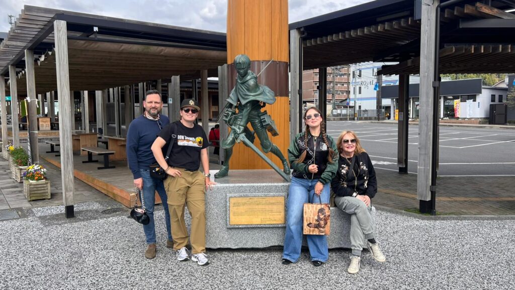 Cuatro personas posan sonrientes alrededor de una estatua de un personaje de anime con espadas, colocada sobre un pedestal de piedra. Dos están de pie a la izquierda y dos sentadas a la derecha del monumento. Llevan ropa casual y gafas de sol. La escena se encuentra en un espacio urbano abierto con estructuras de madera, bancos y una calle al fondo.